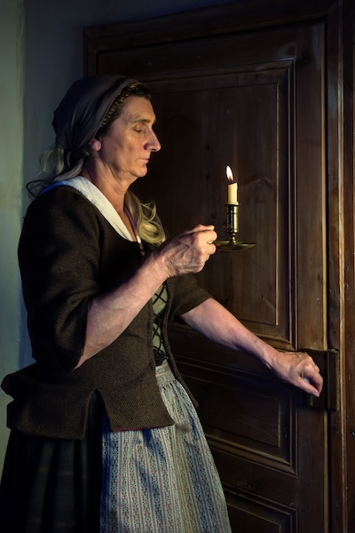 The image displays a woman holding a candle lantern to move from room to room in her house. 