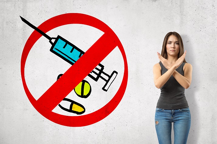 anti-vaxer woman Serious young girl with hands crossed, standing against wall with picture of syringe and some meds enclosed in red prohibition sign.
