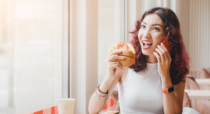 A woman eats a hamburger in a fast food cafe and talks to a friend on the phone