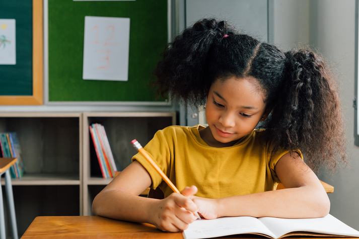 African American school girl sitting in school writing in note book with pencil