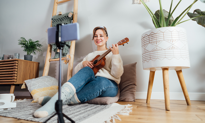 Young Woman playing ukulele guitar, recording music video on phone in modern Scandi interior home.