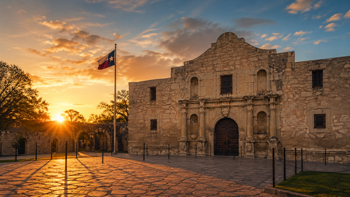 A wide, rectangular image of the Alamo mission at sunrise, with long shadows and a quiet, dramatic sky.