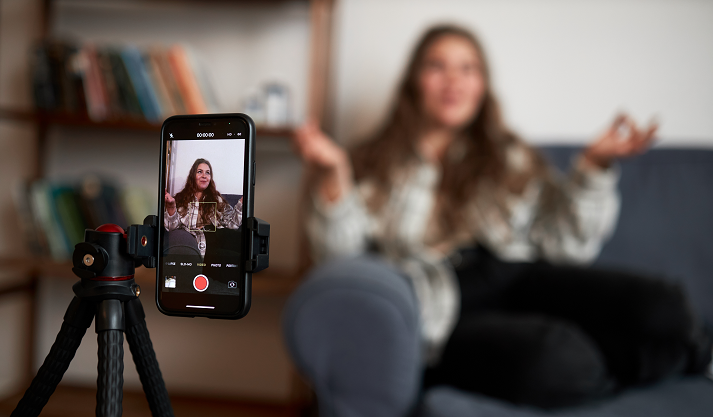 Selective focus on phone on tripod recording vlogger sitting on couch gesticulating and communicating with her followers, livestreaming for her blog in social media.